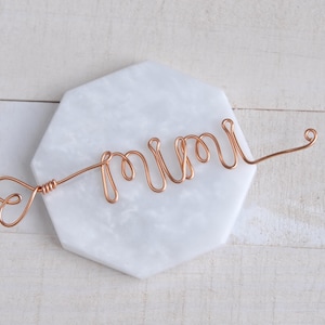 May include: A copper wire sculpture of the word "mum" with a heart at the beginning of the wire. The sculpture is on a white marble surface.