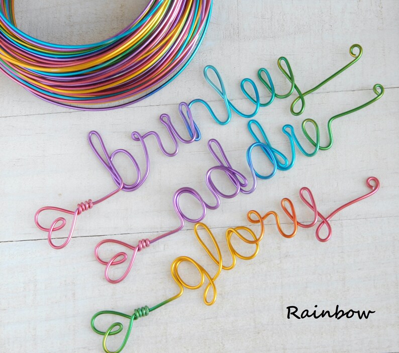 May include: A coil of multi-colored craft wire and three wire names shaped in cursive script: Brinley in purple, Addie in blue, and Glory in yellow-orange. Each name features a small heart-shaped loop at the end. The text Rainbow is visible.