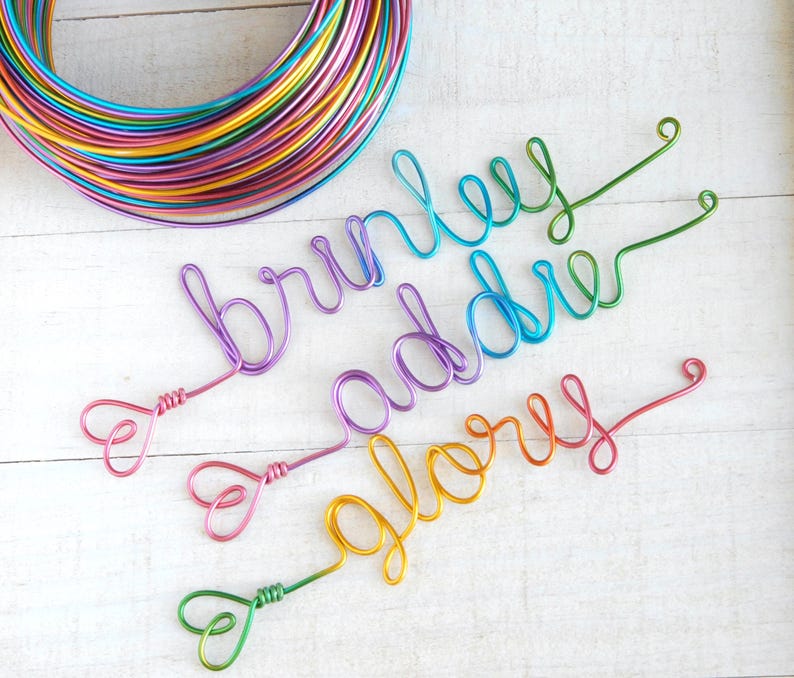 May include: Three custom wire names shaped into cursive script reading Brinley, Addie, and Glory. Each name is crafted from multicolored metallic wire, featuring a small heart-shaped loop at the beginning and a coiled end. A large coil of rainbow wire sits above.