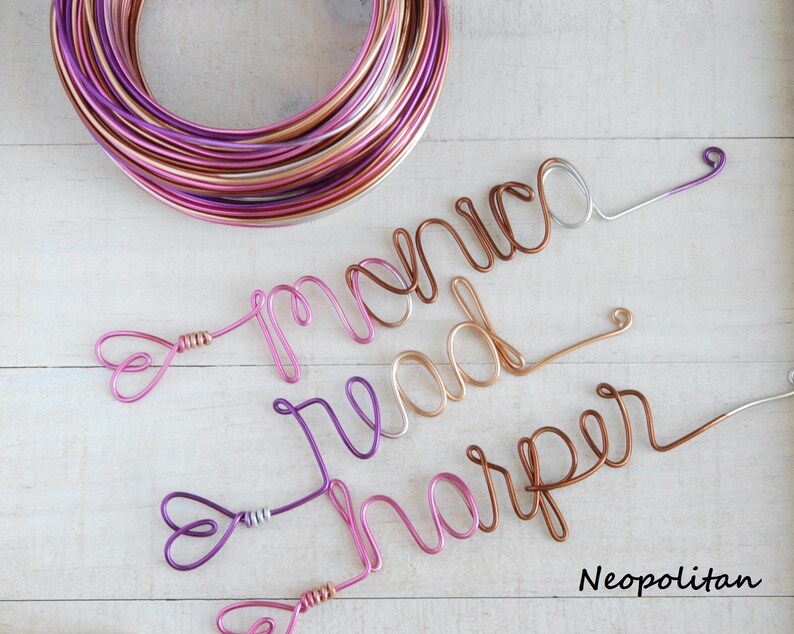 May include: A coil of multi-colored metallic wire in shades of pink, copper, and silver, alongside three wire-formed names: monica, read, and harper. Each name is crafted from continuous wire in varying metallic tones with heart-shaped ends.