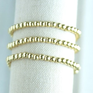 May include: Three gold beaded bracelets wrapped around a white fabric cylinder. The bracelets are made of small, round, gold beads. The fabric has a textured appearance.