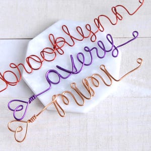 May include: Three custom wire names crafted in cursive script. The top name is red, the middle name is purple, and the bottom name is copper. Each wire name features a small heart shape at the end, with the names reading Brooklyn, Avery, and Mimi.