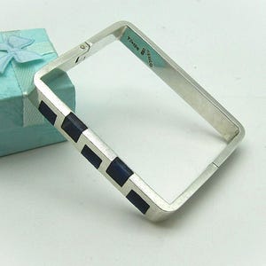 May include: A silver square bangle bracelet with inlaid dark blue rectangular stones. The bracelet is next to a light blue gift box with a bow.
