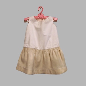May include: A sleeveless dress featuring a solid white bodice and a contrasting beige skirt with a gathered waistline. The hem of the skirt is finished with a decorative white stitched pattern. The dress is displayed on a pink plastic hanger.