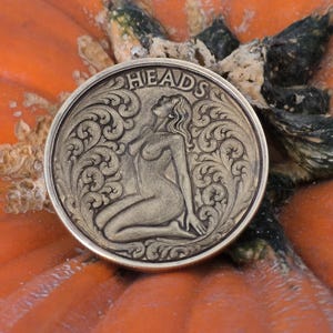 May include: A circular coin with the word "HEADS" at the top, featuring an engraved design of a kneeling woman. The coin has an antique finish and is set against an orange background. The coin is surrounded by ornate scrollwork.