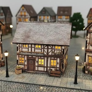 May include: A model village scene with several illuminated houses. The houses have brown and white timber-frame designs, lit windows and doors, and are set on a cobblestone street with street lamps.