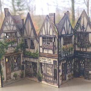 May include: A decorative folding screen featuring a detailed illustration of a row of Tudor-style buildings. The scene includes storefronts labeled Cream Tea & Coffee Cafe, Florist Worth, and Candles, with intricate timber framing and floral accents.