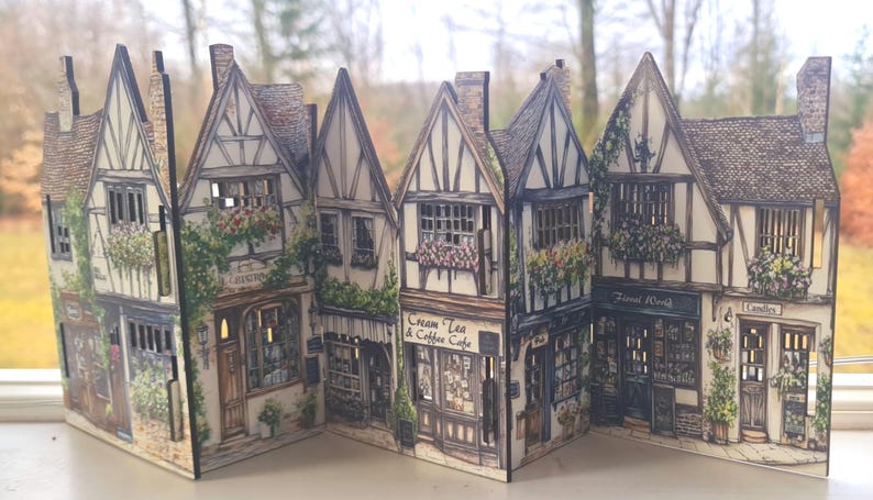 May include: A decorative folding screen featuring a detailed illustration of a row of Tudor-style buildings. The scene includes storefronts labeled Cream Tea & Coffee Cafe, Florist Worth, and Candles, with intricate timber framing and floral accents.