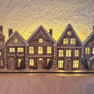 May include: A decorative wooden gingerbread house display featuring five illuminated houses with intricate icing details. The houses have lit windows and are arranged on a wooden base, creating a warm, festive ambiance. The houses are brown with white icing details.