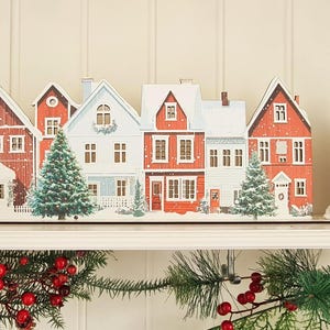 Op de afbeelding: Een decoratief houten dorpsdisplay met een rij rode en witte huizen met besneeuwde daken, groene dennenbomen en ramen. Daarnaast staat een klein wit stoffen zakje met een rood geborduurd hart en een grijze metalen koffiebus.