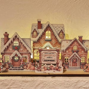 May include: A decorative holiday village display featuring five illuminated gingerbread-style houses with snow-dusted roofs, a central sign reading "Bake Shop Gingerbread Baking Co.", a gingerbread man figure, a nutcracker, and a Christmas tree.