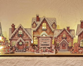 Può includere: Un villaggio natalizio decorativo con cinque case illuminate in stile pan di zenzero con tetti innevati, un cartello centrale con la scritta "Bake Shop Gingerbread Baking Co.", un omino di pan di zenzero, uno schiaccianoci e un albero di Natale.