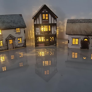 May include: Three illuminated miniature houses with detailed facades. The houses have different architectural styles, including a cottage with a thatched roof, a stone house, and a timber-framed building. The warm interior lights create a cozy ambiance.