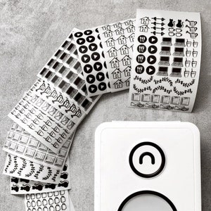 May include: A set of black and white sticker sheets with various icons and designs. The stickers include icons for houses, play buttons, laptops, coffee cups, watering cans, and more. The stickers are arranged in a fan shape on a gray surface.