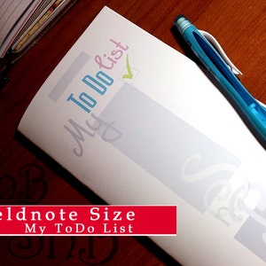 May include: A white to-do list notepad with a blue and green checkmark. The notepad is labeled "My To Do List" in a light blue and pink font. A blue pen is lying on top of the notepad. The notepad is labeled "Fieldnote Size My To Do List" in red.