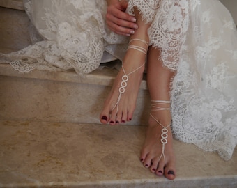 Puede incluir: Un par de sandalias blancas para pies descalzos con múltiples adornos circulares. Las sandalias se llevan en los pies de una mujer, con los dedos pintados de un color rojo oscuro.