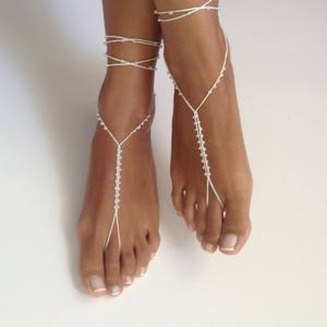 May include: White beaded foot jewelry with a delicate design. The jewelry is made of small white beads strung on a thin white cord. The jewelry is worn on both feet, with the cords wrapping around the ankles and toes.