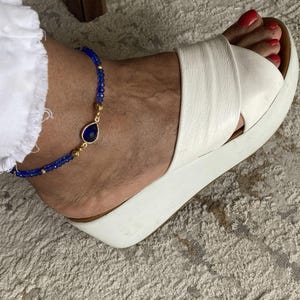 May include: A blue beaded anklet with a teardrop-shaped dark blue stone and gold accents. The anklet is worn on an ankle with a white platform sandal. The person's toenails are painted red.