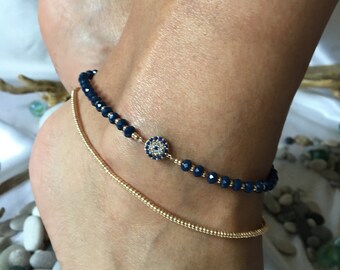 May include: A gold anklet with a blue evil eye charm and a second gold anklet with tiny beads. The anklets are on a person's ankle.