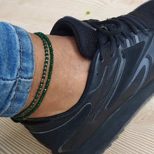 May include: A pair of green beaded anklets on a person's ankle. The anklets are made of small, round beads that are strung together on a thin cord. The anklets are worn on the ankle of a person wearing black trainers and blue jeans.