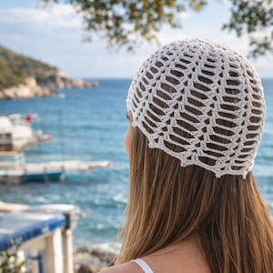 May include: A white, open-weave crochet hat. The hat is round and covers the top of the head. The background shows a view of the ocean and a blue sky. The hat is a light, airy accessory.