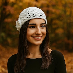 May include: A white crocheted hat with a floral accent. The hat is made of a textured, open-weave material. The background is a blurred view of autumn foliage.
