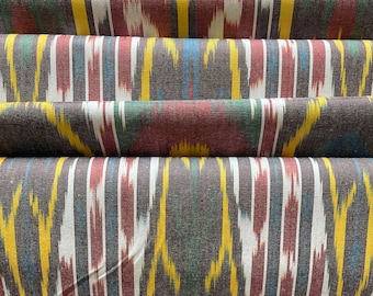May include: Close-up of a folded fabric with a repeating ikat pattern. The design features vertical stripes in shades of brown, yellow, red, green, white, and blue. The fabric has a textured appearance, with visible folds and creases.