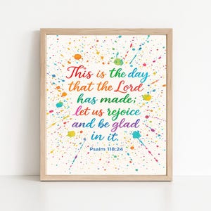 Op de afbeelding: Een ingelijste print met de tekst "This is the day that the Lord has made; let us rejoice and be glad in it. Psalm 118:24." De tekst is in regenboogkleuren, met een achtergrond van kleurrijke verfspatten.