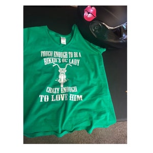 May include: Green t-shirt with a white graphic that says "Tough enough to be a biker's ol' lady, crazy enough to love him". The graphic also includes a motorcycle.