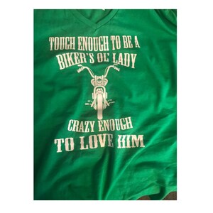 May include: Green t-shirt with white text that reads "Tough enough to be a biker's ol' lady. Crazy enough to love him."  A white silhouette of a motorcycle is between the two lines of text.