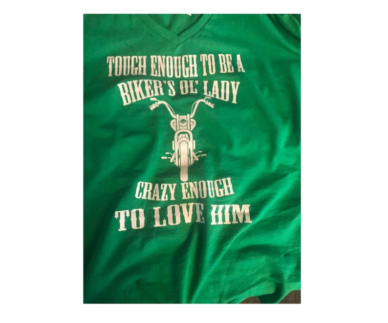 May include: Green t-shirt with white text that reads "Tough enough to be a biker's ol' lady. Crazy enough to love him."  A white silhouette of a motorcycle is between the two lines of text.