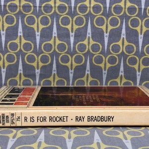 May include: A vintage paperback book titled "R is for Rocket" by Ray Bradbury. The book cover features a colorful illustration of a rocket ship launching into space. The book is published by Bantam Pathfinder Edition and costs 75 cents.