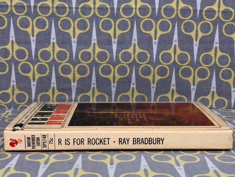 May include: A vintage paperback book titled "R is for Rocket" by Ray Bradbury. The book cover features a colorful illustration of a rocket ship launching into space. The book is published by Bantam Pathfinder Edition and costs 75 cents.