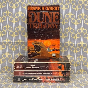May include: A stack of Frank Herbert's "Dune" books. The top book is "The Complete Dune Trilogy" with a sunset and desert landscape illustration. Below are "Dune", "Dune Messiah", and "Children of Dune". The books are in shades of brown and orange.