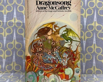 以下が含まれることがあります： アン・マキャフリー著のペーパーバック『Dragonsong』。表紙には、小さな黄金のドラゴンを抱えた人物が中央に描かれており、その周囲を緑、金、茶色の色調の複数のドラゴンが渦巻く背景の中で囲んでいるイラストが施されています。