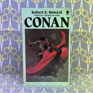 May include: A paperback book cover featuring a dark fantasy illustration of a muscular man in a red cape fighting a monstrous creature. The book title is "CONAN" and the author is Robert E. Howard.