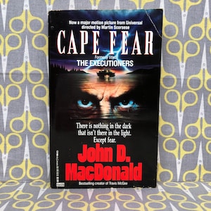 May include: A paperback book cover with a black background and a close-up of a man's face with piercing blue eyes. The title of the book is "Cape Fear" and the author is "John D. MacDonald". The book is a novel based on the movie of the same name directed by Martin Scorsese.