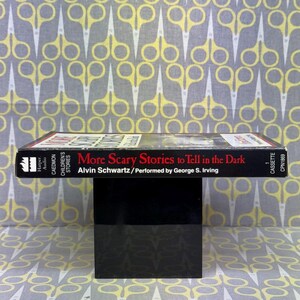 Puede incluir: Una cinta de cassette negra con el t&iacute;tulo "More Scary Stories to Tell in the Dark" de Alvin Schwartz, interpretada por George S. Irving. El texto es blanco y rojo. El fondo es una tela estampada con tijeras amarillas.