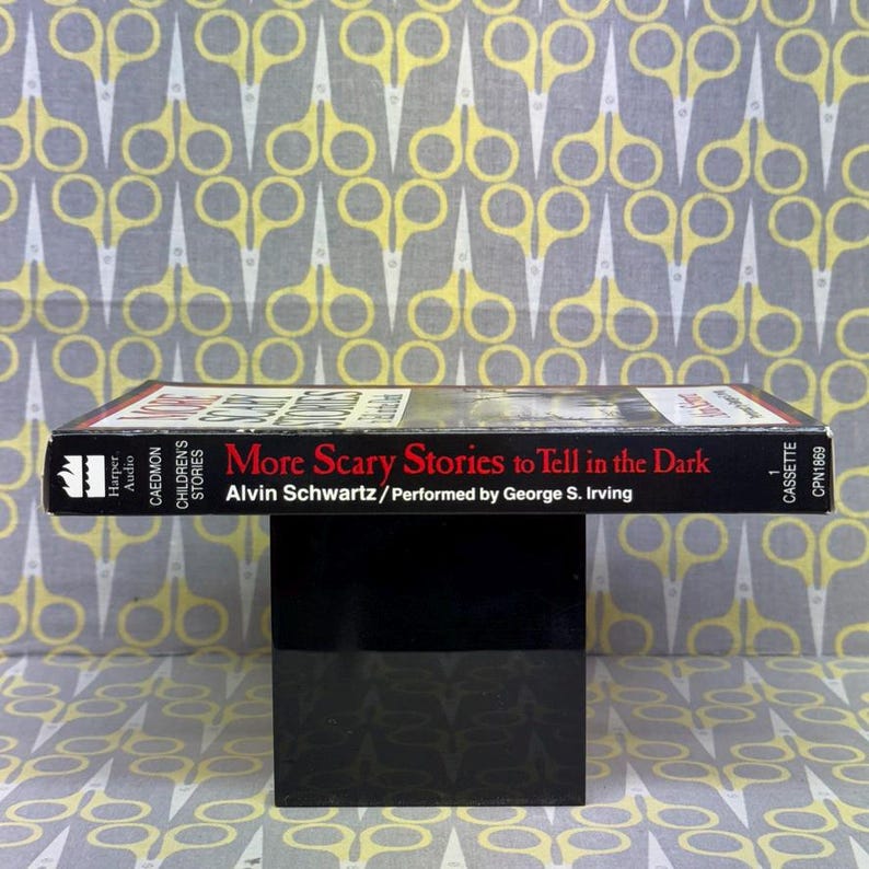 Puede incluir: Una cinta de cassette negra con el t&iacute;tulo "More Scary Stories to Tell in the Dark" de Alvin Schwartz, interpretada por George S. Irving. El texto es blanco y rojo. El fondo es una tela estampada con tijeras amarillas.