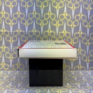 Puede incluir: Una caja rectangular con la parte superior blanca y ribete rojo se encuentra sobre un soporte negro. El fondo presenta un patr&oacute;n repetido de tijeras amarillas y blancas sobre un fondo gris. La caja parece contener material impreso.