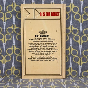 May include: A yellow and brown paperback book with the title "R is for Rocket" and the author "Ray Bradbury". The back cover features a description of the book, which includes the text "The Spellbinding Power of Ray Bradbury".