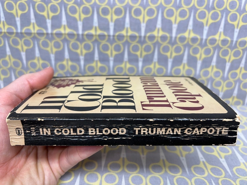 May include: A black paperback book with the title "In Cold Blood" by Truman Capote. The book is worn and has a few scratches on the cover.