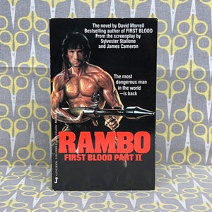 May include: A paperback book cover for the novel "Rambo: First Blood Part II" by David Morrell. The cover features a photo of Sylvester Stallone as John Rambo, holding a rocket launcher. The text on the cover reads "The most dangerous man in the world - is back".