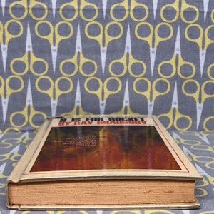 May include: A vintage hardcover book titled "I Is for Rocket" by Ray Bradbury. The cover features a brown and orange abstract design.