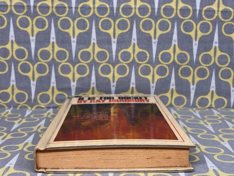 May include: A vintage hardcover book titled "I Is for Rocket" by Ray Bradbury. The cover features a brown and orange abstract design.