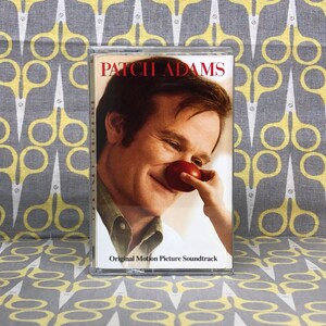 Puede incluir: Una cinta de casete con una imagen de Robin Williams en la portada. El título de la cinta es "Patch Adams" y el texto "Original Motion Picture Soundtrack" está debajo del título.