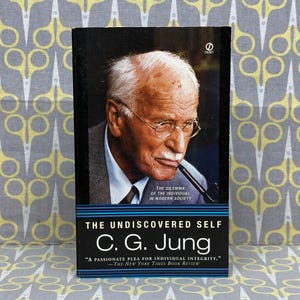 以下が含まれることがあります： C.G.ユング著「The Undiscovered Self」というタイトルのペーパーバック。表紙には、眼鏡とパイプを持った老人の肖像画が描かれています。本は青と黒の表紙に白い文字で書かれています。本は「個人の誠実さへの情熱的な嘆願」と説明されています。