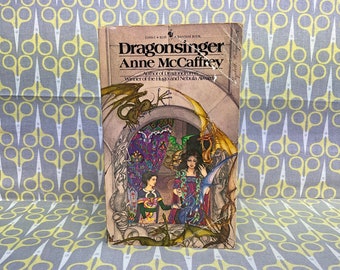 以下が含まれることがあります： 「ドラゴンスインガー」というタイトルのヴィンテージペーパーバックの表紙。アン・マッカフリー著。表紙には、長い黒髪の女性が、後ろにドラゴンがいるドアの入り口に立っているイラストが描かれている。この本はバンタム・ブックである。