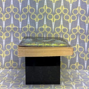 May include: A hardcover book with a green and black cover, resting on a black cube. The book's pages are yellowed with age. The background features a repeating pattern of yellow scissors on a gray background.