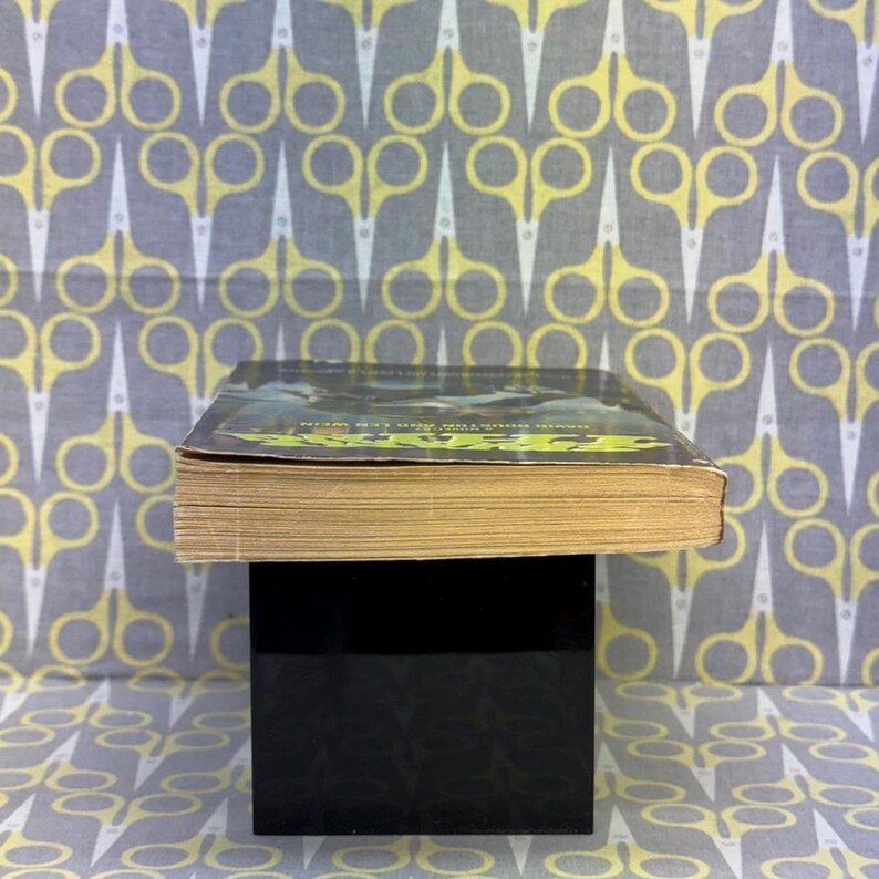 May include: A hardcover book with a green and black cover, resting on a black cube. The book's pages are yellowed with age. The background features a repeating pattern of yellow scissors on a gray background.